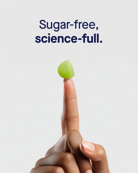 A close-up photograph of a single green vitamin gummy held between two fingers against a clean, neutral background.